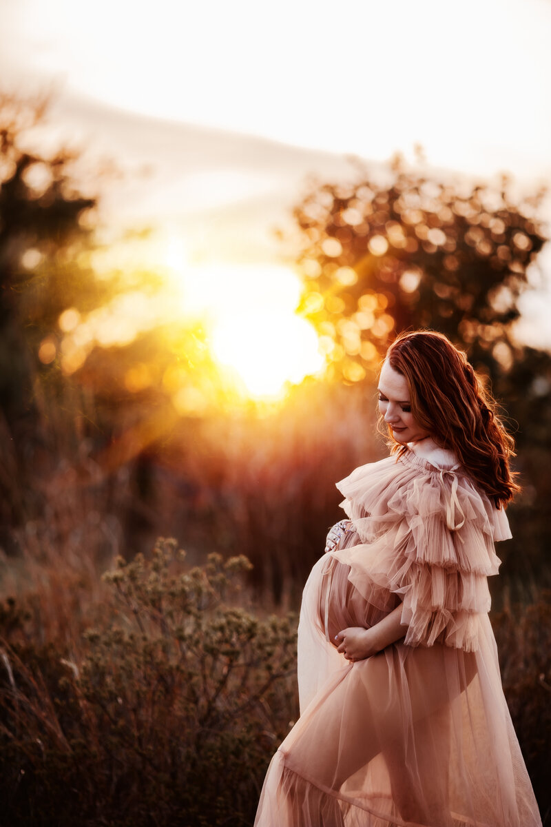 A pregnant woman standing looking down at her belly with a beautiful golden sunset light in the background